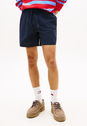 Moški, ki nosi temno modre kratke hlače, bele nogavice z logotipom, bež čipkaste čevlje in rdeče-modro črtasto majico z dolgimi rokavi, z rokami v žepih.