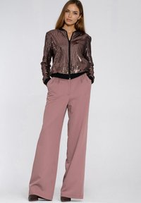 Błyszcząca kurtka bomberka w kolorze brązowym z czarnymi ściągaczami, zestawiona z szerokimi różowymi spodniami. Oba elementy charakteryzują się luźnym krojem.