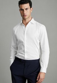 Weißes Langarm-Hemd mit Knöpfen, klassischem Kragen und glattem Stoff, kombiniert mit dunkelblauen Hosen. Minimalistisches Design ohne Muster.
