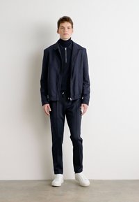 Chaqueta de navy y cuello alto, combinada con pantalones a juego. Zapatillas blancas. Diseño minimalista, textura suave y un sutil brillo en la tela.