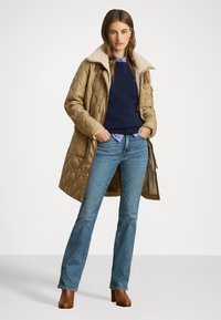 Kvādrainā krēmbaltā virsjakā ar aitas vilnas apkakli, tumši zilā džemperī, gaišzila krekli, un zili džinsi; apģērbu papildina brūni apavi ar papēžiem.