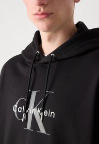 Crna dukserica s kapuljačom, s bijelim logotipom "Calvin Klein" i sivim "K" sprijeda. Sadrži crne vezice i ležeran kroj.