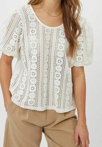 Blusa bianca in pizzo con scollatura rotonda e maniche a sbuffo, caratterizzata da un motivo floreale e dettagli di ricamo aperto, abbinata a pantaloni beige.