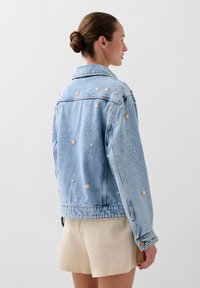 Giacca di jeans azzurro chiaro con decorazioni metalliche, vestibilità oversize e design del colletto classico. Tessuto testurizzato con chiusura a bottoni.