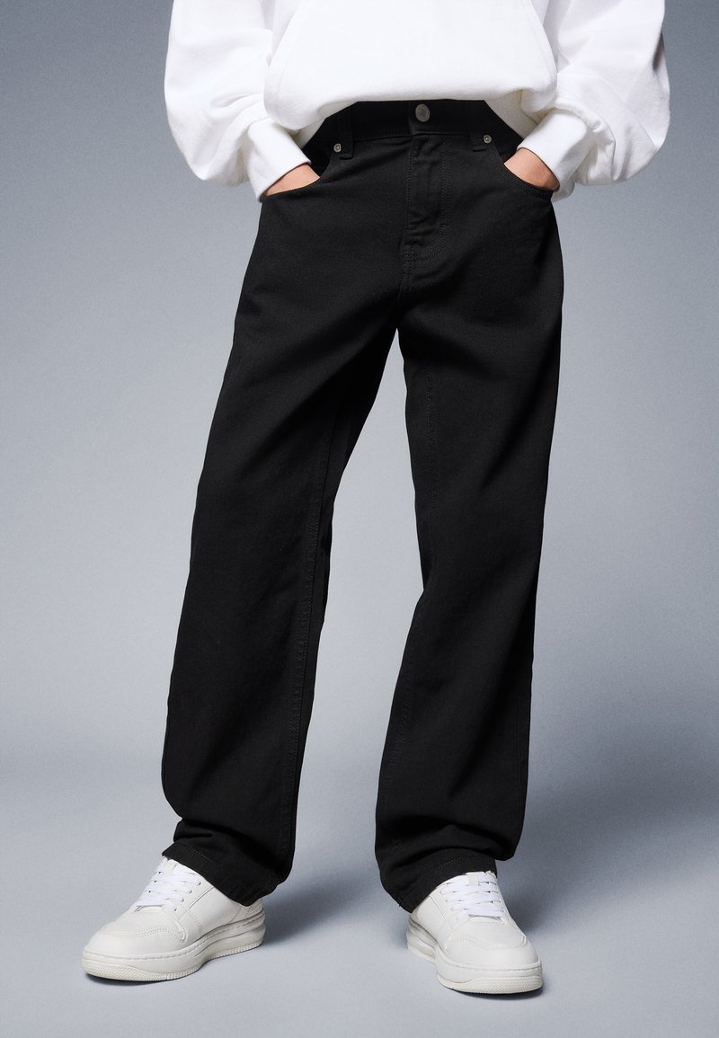Black denim jeans with a loose fit, high waist, and front pockets. Paired with white sneakers, on a neutral background.