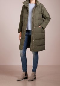 Olivgrüner Puffer-Mantel mit hohem Kragen, Reißverschluss vorne und Seitentaschen. Getragen über einem hellblauen Pullover und enganliegenden Jeans, kombiniert mit grauen Ankle Boots.