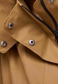 Bruine waterdichte jas met een gestructureerde kraag, zilveren drukknoopsluitingen en een centrale rits. Glad stofoppervlak.