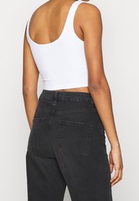 Mujer con un top corto sin mangas blanco y jeans negros de cintura alta, vista desde atrás contra un fondo claro y liso.