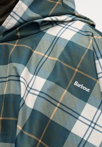 Ύφασμα με καρό μοτίβο σε πράσινο, λευκό και πορτοκαλί, με το λογότυπο της Barbour κεντημένο σε διπλωμένο μπουφάν.