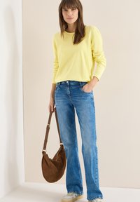 Femme portant un haut à manches longues jaune et un jean bleu, tenant un sac à bandoulière marron, debout devant un fond beige uni.