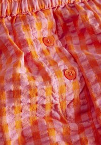Detailný záber na látku v ružovom a oranžovom kockovaní s dvoma oranžovými gombíkmi a zhromaždeným elastickým pásom.