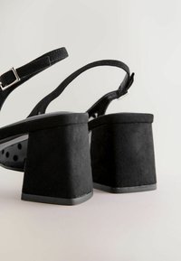 Mustat mokkanahkaiset korkokengät, joissa on paksu, neliskulmainen korko. Niissä on ohut nilkkahihna metallisella soljella ja pilkkukuosinen sisäpohja.