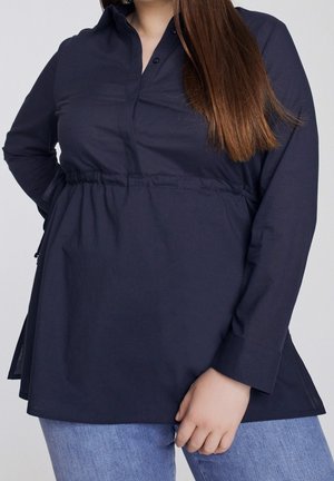 Kvinde iført en langærmet mørkeblå tunika med en indsnøret talje og lyseblå jeans, vist fra skuldre til hofter.