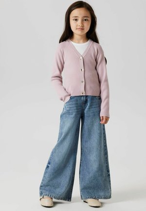 Lichtroze geribde cardigan met zes glanzende knopen, gedragen over een witte top, in combinatie met wijde blauwe jeans en beige schoenen.