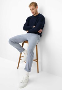 Man in marineblauwe trui en lichtblauwe broek zit op een houten kruk, armen gekruist, tegen een witte muur en vloer.