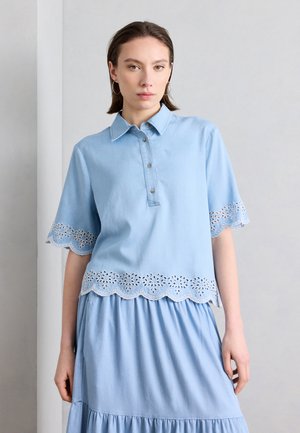 Młoda kobieta ubrana w jasnoniebieską bluzkę z kwiatową koronką ażurową na rękawach i dole, w połączeniu z pasującą jasnoniebieską spódnicą.