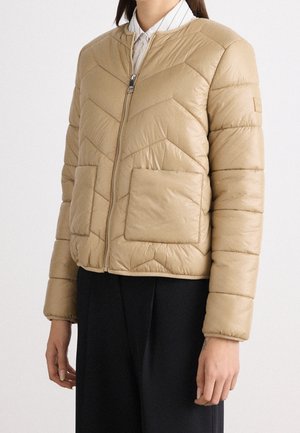 Beige gewatteerde jas met ritssluiting en grote zakken aan de voorkant, gedragen over een wit overhemd met kraag en zwarte broek.