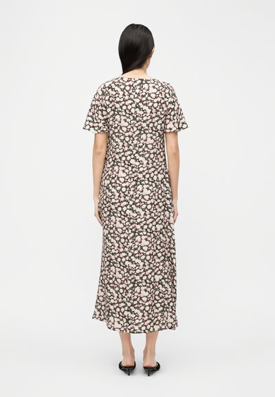 Γυναίκα που φοράει μαύρο μεσαίο φόρεμα με ρόζ και λευκά λουλουδένια σχέδια, κοντά μανίκια και μαύρα παπούτσια με χαμηλό τακούνι, στέκεται με την πλάτη στραμμένη.