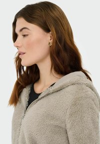 Sweat à capuche gris clair et duveteux avec une fermeture éclair à l'avant et une texture douce, porté par-dessus un haut noir. Des boucles d'oreilles clous en forme de fleur dorée ajoutent une touche de détail.