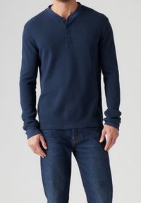 Navyblaues, geripptes Henley-Shirt mit langen Ärmeln, Rundhalsausschnitt, Dreiknopfleiste und angesetzten Bündchen. Kombiniert mit dunkelblauen Jeans.