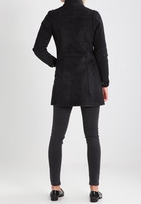 Vrouw die staat, gekleed in een strakke zwarte lange jas, slanke zwarte broek en zwarte instappers, van achteren weergegeven op een eenvoudige achtergrond.