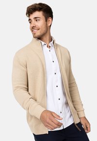 Beige gebreide cardigan met ribbelmanchetten en -zoom, voorzien van een ritsluiting. Draagt over een wit overhemd met knopen. Licht, gestructureerde stof.