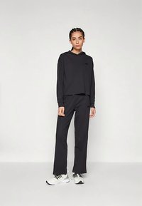 Μαύρο φούτερ με κουκούλα σε crop σχεδίαση και κεντημένο λογότυπο, σε συνδυασμό με μαύρο παντελόνι που ταιριάζει και λευκά αθλητικά παπούτσια.