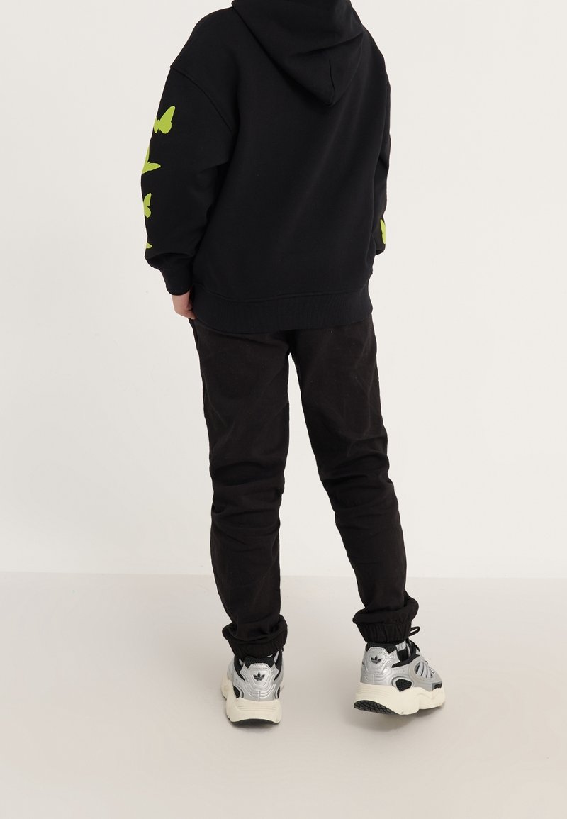 Personne portant un sweat à capuche noir avec des papillons verts sur les manches, un pantalon noir et des baskets argentées, debout devant un fond clair uni.