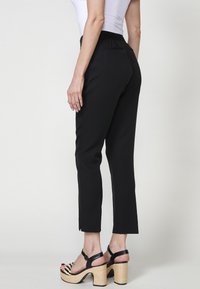 Pantalons noirs ajustés avec une taille élastique, longueur cropped et détails de poche arrière subtils, associés à des sandales à talons hauts noires et tressées.