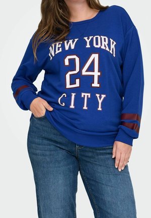 Kobieta w niebieskiej bluzie z "New York City" i numerem 24, w niebieskich dżinsach, z ręką w kieszeni, na jednolitym tle.