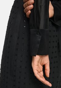 Schwarzes Kleid aus strukturiertem Stoff mit einem Gittermuster. Der Ärmel ist aus Satin mit einem Knopfdetail. Eine Hand hält den Ärmel.