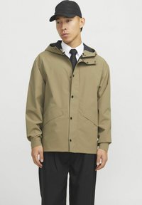 Khaki waterdichte jas met een capuchon, drukknoopsluiting en twee voorkruiszakken. Draagt over een wit shirt en zwarte stropdas, in combinatie met zwarte broek.