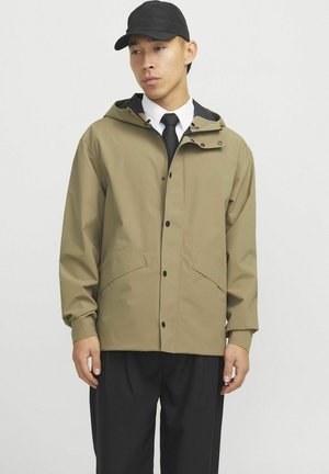 Veste imperméable kaki avec une capuche, des boutons-pression et deux poches avant. Portée sur une chemise blanche et une cravate noire, associée à un pantalon noir.