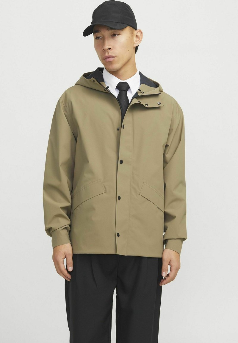 Khaki waterdichte jas met een capuchon, drukknoopsluiting en twee voorkruiszakken. Draagt over een wit shirt en zwarte stropdas, in combinatie met zwarte broek.