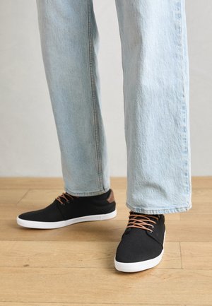Zapatillas de lona negras con detalles marrones, suelas de goma blanca y cordones marrones, combinadas con jeans rectos de color azul claro. Fondo de suelo de madera.