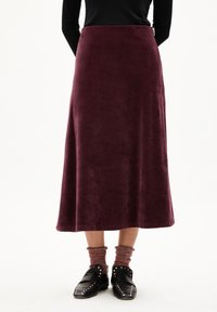 Femme portant une longue jupe bordeaux en tissu texturé, des chaussettes rouges marbrées et des chaussures à enfiler noires avec clous, se tenant devant un fond blanc.