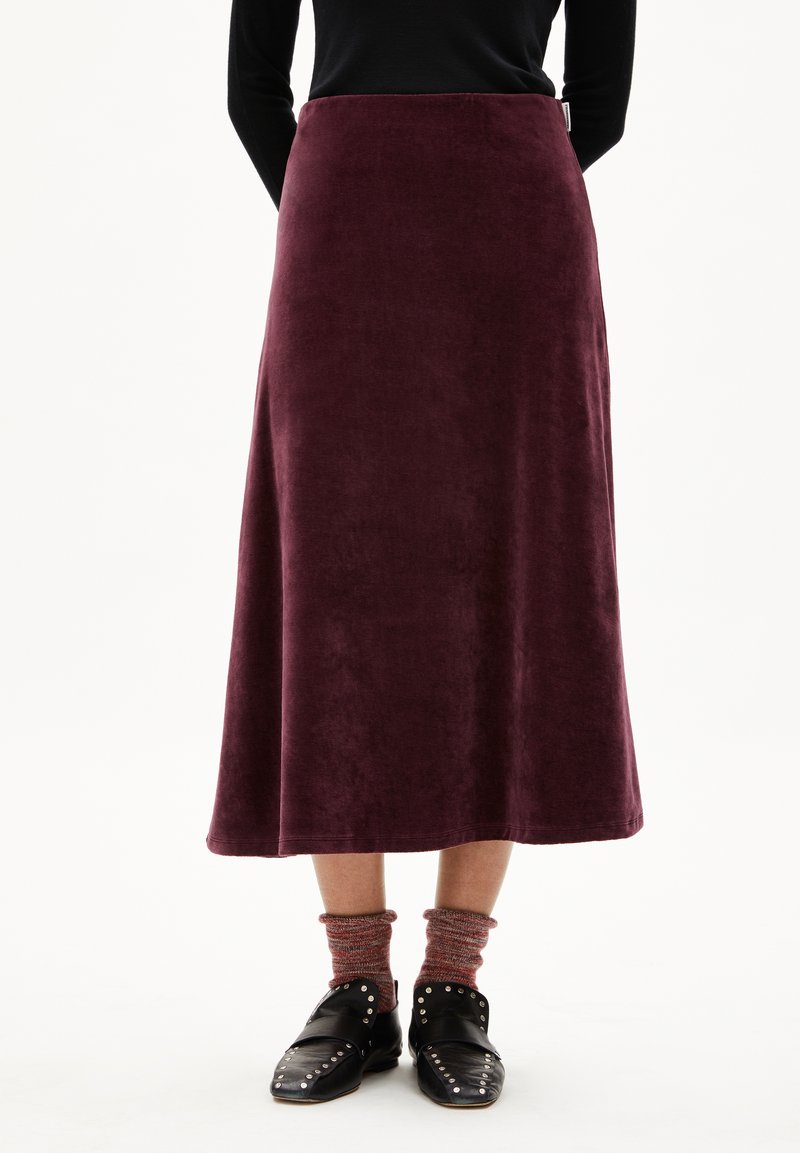 Femme portant une longue jupe bordeaux en tissu texturé, des chaussettes rouges marbrées et des chaussures à enfiler noires avec clous, se tenant devant un fond blanc.