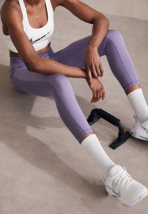 Leggings deportivos morados y un top corto blanco, acompañados de calcetines blancos con canalé y zapatillas blancas, descansando junto a un soporte de flexiones negro.