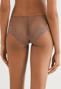 Kvinna som bär genomskinligt brunt spetsunderkläder, sedd bakifrån mot en enkel ljus bakgrund.