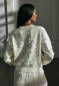Cremefarvet sweater med pailletdetaljer, tekstureret stof og ribbede manchetter. Har en løs pasform og er lagt over en fluffy hvid nederdel.