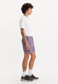 Tricou alb cu mânecă scurtă asociat cu pantaloni scurți ușor violet. Băiat purtând adidași gri și maro cu șosete albe, surprins din lateral.
