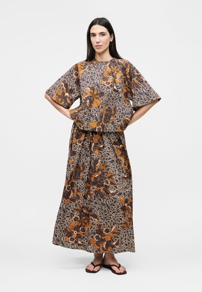 Γυναίκα με μακριά σκούρα μαλλιά που φοράει ένα χαλαρό τοπ με λουλουδάτο σχέδιο και μακριά φούστα στο ίδιο μοτίβο, στέκεται με τα χέρια στη μέση και σανδάλια.