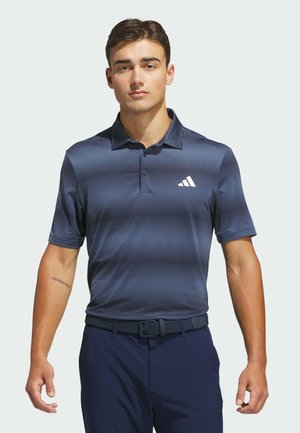 Mężczyzna ubrany w granatową koszulkę polo w paski z białym logo, ciemnym paskiem i ciemnymi spodniami, stojący na tle jednolitego jasnego tła.