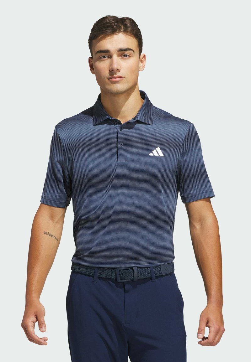Homme portant un polo rayé bleu marine avec un logo blanc, une ceinture sombre et un pantalon sombre, debout devant un fond clair uni.