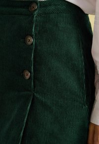 Grüne Cordrock mit vertikalen Rippungen, ausgestattet mit braunen Knopfverschlüssen an der Seite und einem Schlitz für zusätzliche Details.