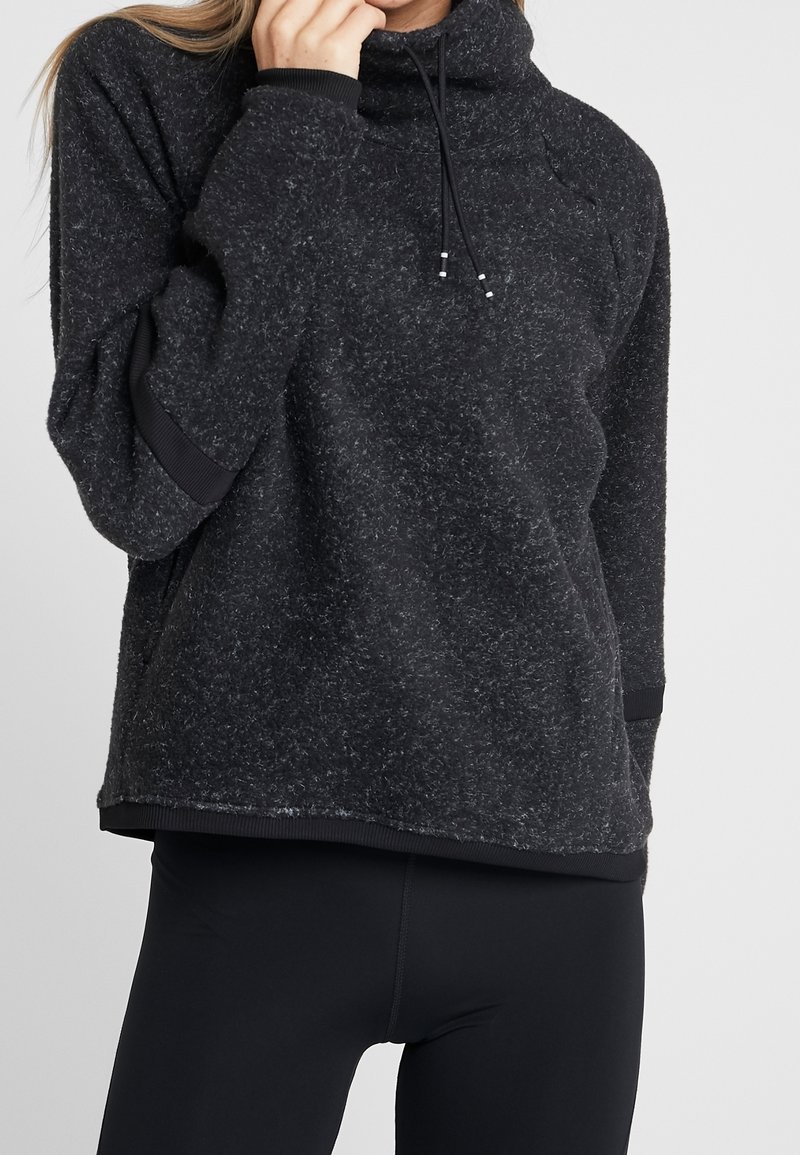 Femme portant un pull polaire gris foncé avec col montant et cordons, associé à des leggings noirs sur un fond uni.