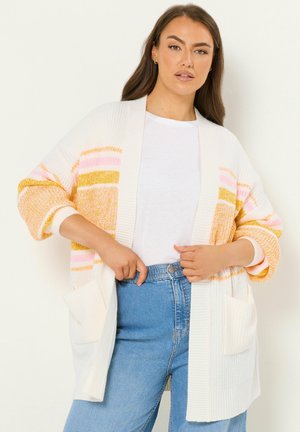 Femme portant un cardigan blanc ouvert avec des rayures jaunes et roses, associé à un t-shirt blanc et un jean bleu taille haute.