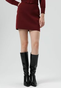 Mini-jupe bordeaux en tissu texturé associée à des bottes en cuir noir montantes jusqu'au genou, avec un bout pointu et une finition brillante.