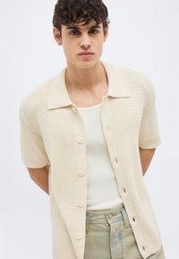 Νεαρός άνδρας με σγουρά μαλλιά, φοράει μια κρεμ κοντομάνικη πλεκτή πουκάμισο με κουμπιά πάνω από λευκό αμάνικο μπλουζάκι και ανοιχτόχρωμο μπλε τζιν.