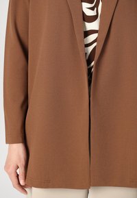 Hnědý látkový blejzr s přiléhavým střihem, obsahující zapínání na jeden knoflík a dlouhé rukávy, přes vzorovaný top.